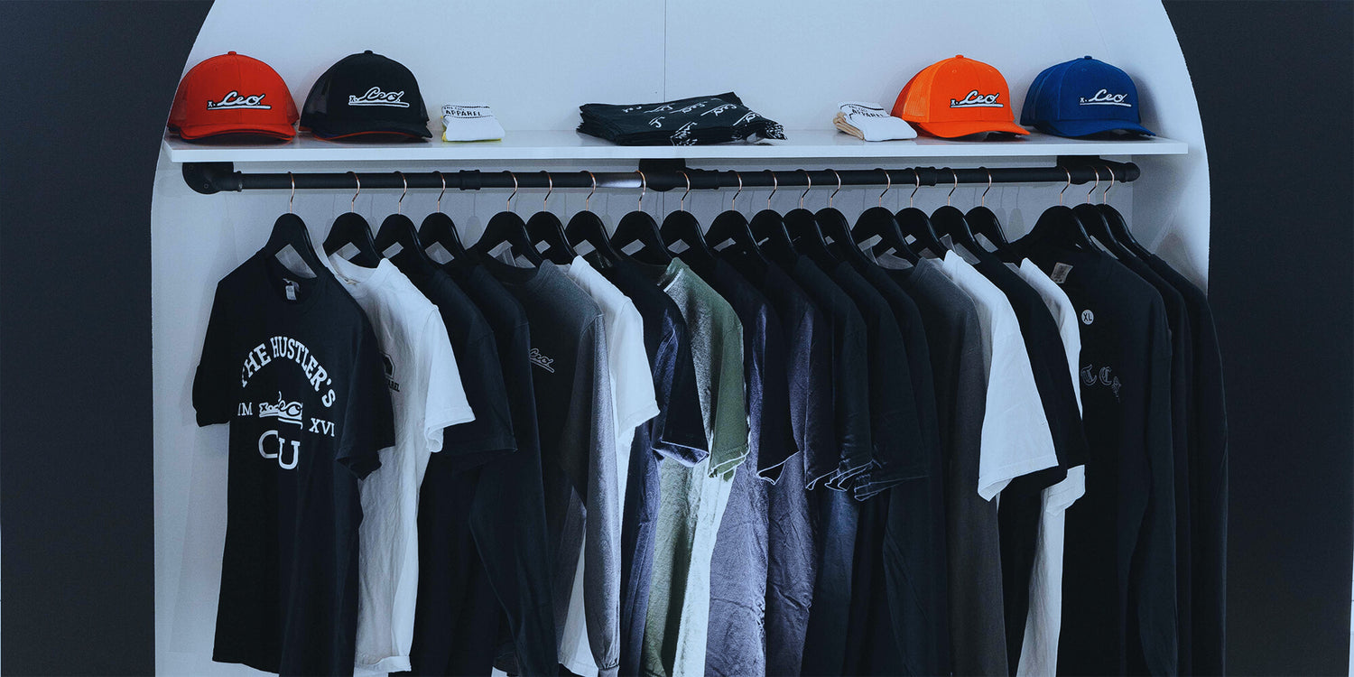 Apparel rack featuring The CEO. Apparel branded hats and a variety of signature t-shirts in black, white, and earth tones displayed in a modern retail setup.